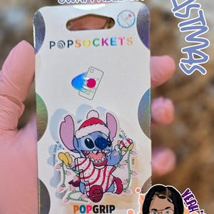 May include: A blue Stitch character wearing a red and white striped shirt and a Santa hat. The character is holding a string of Christmas lights. The image is for a phone grip, a PopSocket, and says "Stitch vs Christmas" and "Swappable Top".