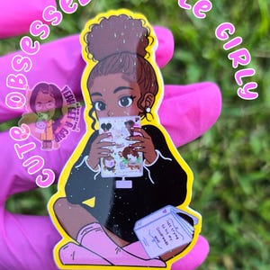May include: A yellow sticker with a cartoon illustration of a Black girl holding a phone. The sticker has the words "CUTE OBSESSED KINDLE GIRLY" in a pink, handwritten font. The girl is wearing a black top and pink socks. A book with the text "Just trying to hit my Goodreads goal" is also visible.