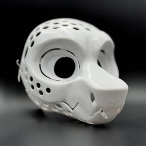 May include: A white, 3D-printed mask with a stylized animal face. The mask features large eye openings, a snout, and numerous small holes for ventilation. The design includes a textured surface and is set against a dark background.