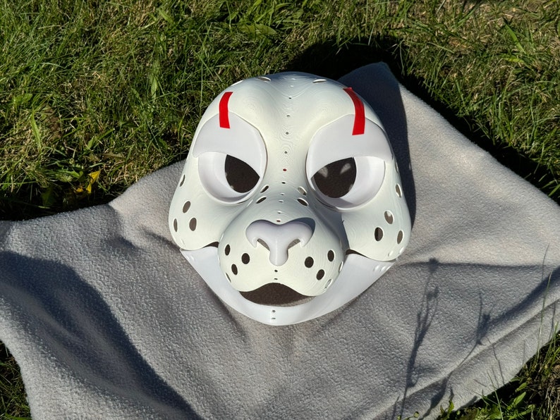 Op de afbeelding: Witte harige dierenmasker met rode accenten en zwarte ooggaten. Het masker heeft een witte neus en een witte mond.
