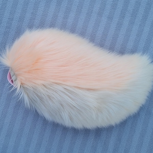 Könnte beinhalten: Ein flauschiger, zweifarbiger Kunstfellschwanz in Pfirsich und Weiß, mit einer rosa Schlaufe am Ansatz. Der Schwanz liegt auf einer blauen, strukturierten Oberfläche und zeigt seine weiche Textur und den Farbkontrast.