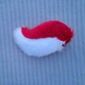 Könnte beinhalten: Ein zweifarbiges, plüschiges Schwanz-Requisit mit einem leuchtend roten Oberteil und einem flauschigen weißen Unterteil. Der Schwanz ist gebogen und hat einen braunen Befestigungsteil. Das Kunstfell wirkt weich und strukturiert.