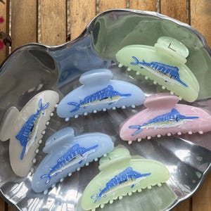 May include: A collection of five translucent hair claw clips in pastel shades of light green, pale blue, and soft pink. Each clip features a detailed graphic of a blue marlin fish on the outer surface, complete with a long bill and fins.