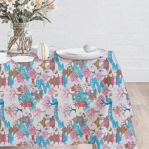 May include: A white tablecloth with a colorful Japanese-inspired pattern featuring pagodas, cherry blossoms, and stylized trees in shades of pink, blue, and green. The table is set with white plates, cups, and a vase of flowers.