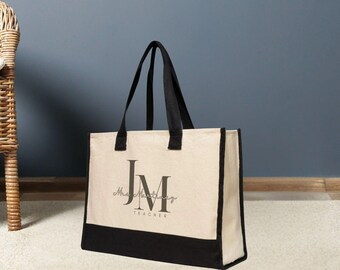May include: A beige tote bag with black accents. The bag features black handles and a black bottom panel. The front of the bag displays the initials "JM" in a stylized font, with the word "Teacher" underneath.