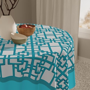 May include: A round tablecloth with a geometric pattern in white and teal covers a table. A teal border trims the edge. A white vase, a small bowl, and a woven basket with fruit sit on the table.