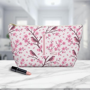 May include: A white cosmetic bag with a pink floral and bird pattern. The bag features a large pink letter "L" and the name "Lillian". A gold zipper and a tube of lipstick are also visible.