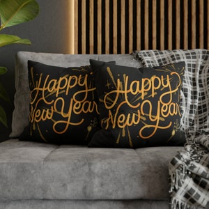 May include: Two black throw pillows with gold lettering that reads "Happy New Year" with gold firework designs. The pillows are on a gray couch with a black and white plaid throw blanket.