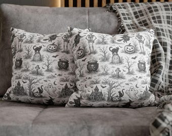 May include: Two square throw pillows with a Halloween-themed pattern. The black and white design features skeletons, pumpkins, black cats, haunted houses, and ghosts. The pillows are resting on a gray sofa, with a plaid blanket in the background.