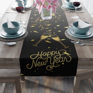 May include: A black table runner featuring a gold-colored graphic print of two clinking champagne flutes, fireworks, and confetti, with the text Happy New Year written in a cursive script font, centered on a wooden dining table with place settings.