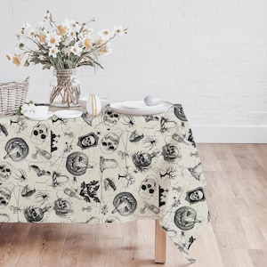 May include: A beige Halloween-themed tablecloth with a repeating pattern of skulls, spiders, bats, pumpkins, and potion bottles in black. The table is set with white plates, a vase of flowers, and decorative eggs.
