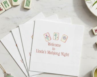 May include: A stack of white paper napkins featuring a printed illustration of three mahjong tiles above the text "Welcome to Linda's Mahjong Night" in a soft pink font. The napkins have a textured border and are arranged in a fan shape.