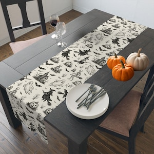 May include: A Halloween-themed table runner with a cream background and black illustrations of ghosts, witches, bats, and pumpkins. The table is set with white plates, forks, and three pumpkins.