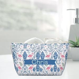 Könnte beinhalten: Eine weiße Kosmetiktasche mit einem Muster aus blauen und rosa Flamingos und Vasen. Die Tasche hat ein blaues Banner mit dem Namen "Chris". Die Tasche hat einen goldenen Reißverschluss.