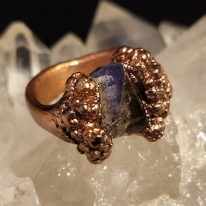May include: A copper ring with a blue gemstone set in a textured copper bezel. The ring is surrounded by small copper beads.