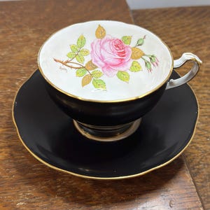 May include: A black teacup and saucer set with a pink rose design. The teacup has a white interior with a gold rim and handle. The saucer is black with a gold rim. The rose is painted in the center of the teacup.