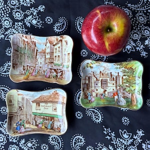 May include: Three small, rectangular decorative trays with curved edges, each featuring a different historical scene. A red apple sits nearby on a black and white paisley patterned cloth. The trays depict street scenes and a castle.
