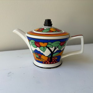 Puede incluir: Tetera de cerámica con tapa, con un diseño colorido de frutas y árboles. La tetera tiene un cuerpo blanco, un asa negra y un pico. La tapa está decorada con frutas rojas, naranjas y verdes. El diseño incluye un árbol, un carro y uvas.