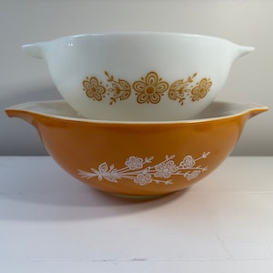May include: Two vintage Pyrex mixing bowls, one white with gold floral pattern and one orange with white floral pattern.