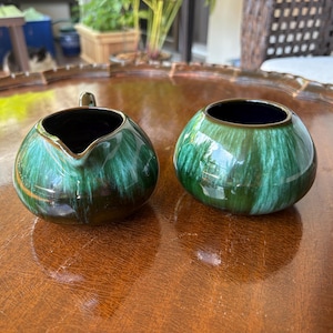 May include: A matching set of ceramic cream and sugar bowls. The bowls are round with a dark green and blue glaze. The creamer has a spout and handle, while the sugar bowl is open. Both have a gold rim.