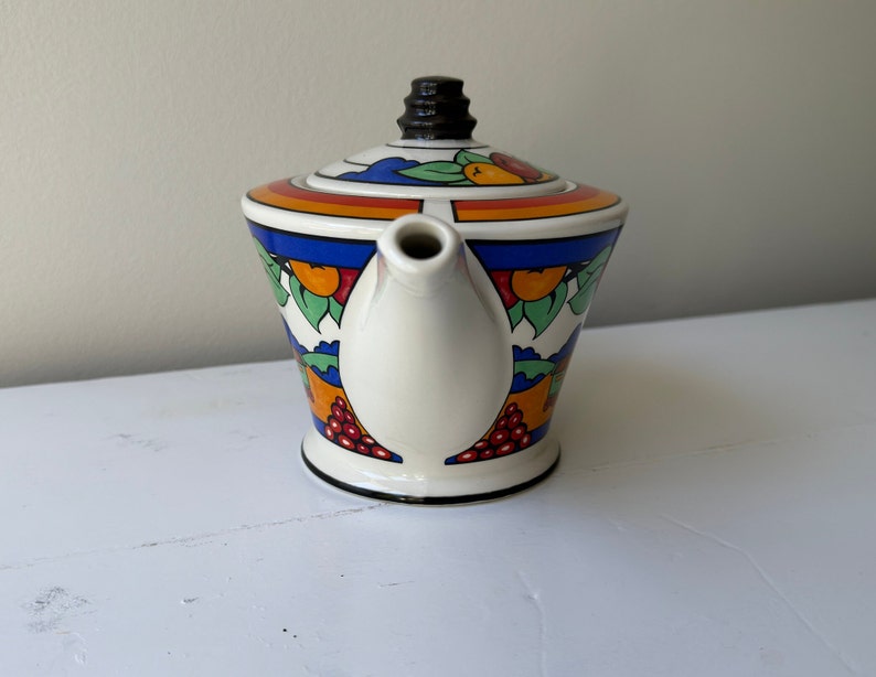Puede incluir: Una tetera de cer&aacute;mica vintage con un colorido dise&ntilde;o de frutas y hojas. La tetera tiene un cuerpo blanco con una base y un asa negras. La tapa tiene un pomo negro y una banda de colores del arco&iacute;ris.