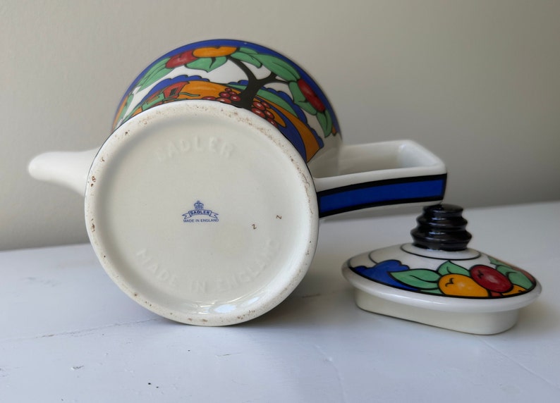 Puede incluir: Una tetera Sadler vintage con un colorido dise&ntilde;o de frutas y &aacute;rboles. La tetera presenta un cuerpo blanco con un asa negra y una franja azul. La tapa est&aacute; a un lado, mostrando el dise&ntilde;o de frutas. La parte inferior de la tetera dice "MADE IN ENGLAND".