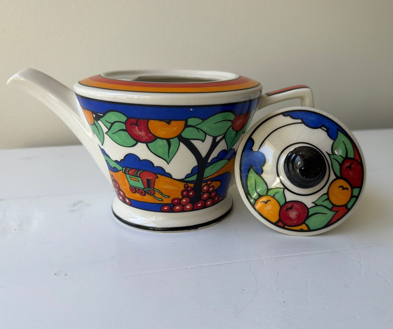 Puede incluir: Tetera de cer&aacute;mica vintage con tapa. La tetera tiene una base blanca con un borde negro, un pico y un asa. El dise&ntilde;o presenta un paisaje con frutas y &aacute;rboles. La tapa tiene un pomo negro y un patr&oacute;n a juego de frutas y hojas.