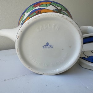 Puede incluir: Tetera de cer&aacute;mica blanca con un dise&ntilde;o de paisaje colorido. La tetera tiene un pico y una tapa con un asa negra. La base est&aacute; estampada con "SADLER" y "MADE IN ENGLAND".