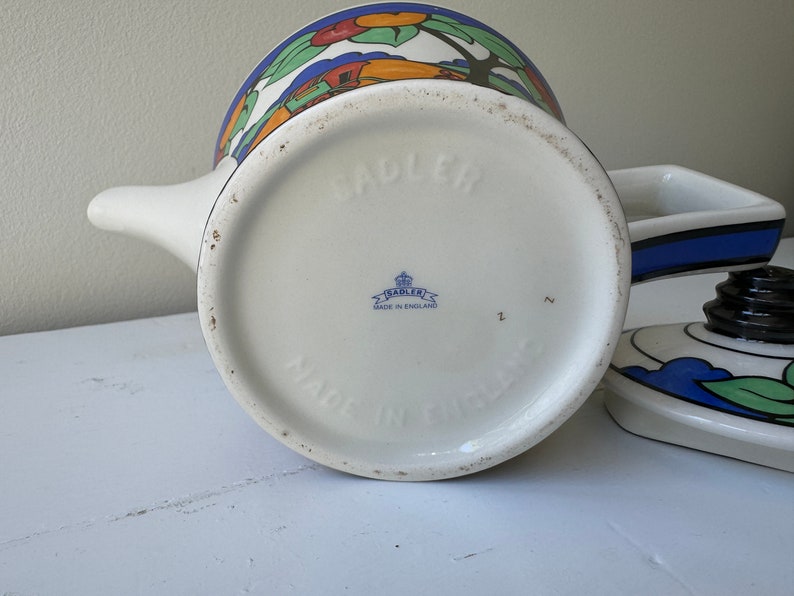 Puede incluir: Tetera de cer&aacute;mica blanca con un dise&ntilde;o de paisaje colorido. La tetera tiene un pico y una tapa con un asa negra. La base est&aacute; estampada con "SADLER" y "MADE IN ENGLAND".