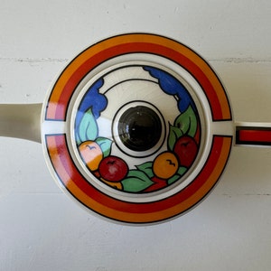 Puede incluir: Tetera de cer&aacute;mica color crema con tapa circular con un dise&ntilde;o vibrante de arco&iacute;ris, frutas y hojas. La tapa tiene un pomo negro. La tetera tiene un pico y un asa con rayas rojas y negras.