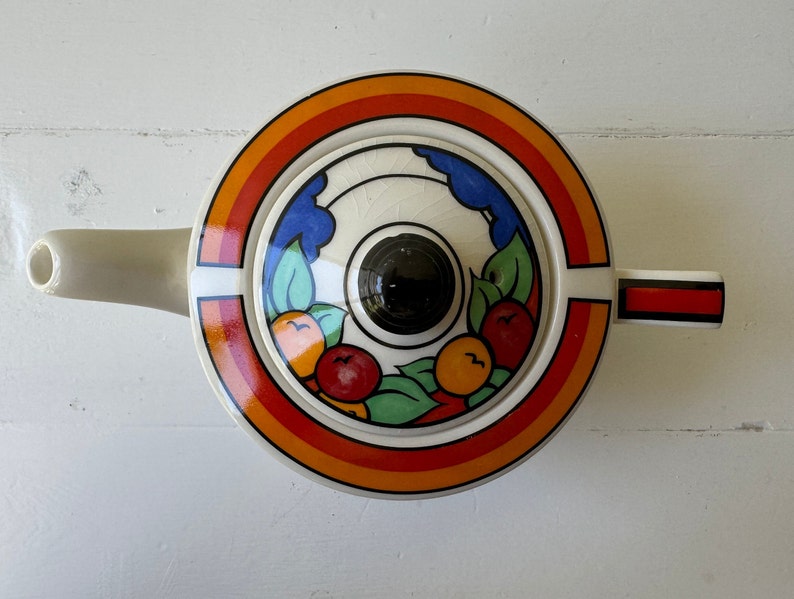 Puede incluir: Tetera de cer&aacute;mica color crema con tapa circular con un dise&ntilde;o vibrante de arco&iacute;ris, frutas y hojas. La tapa tiene un pomo negro. La tetera tiene un pico y un asa con rayas rojas y negras.