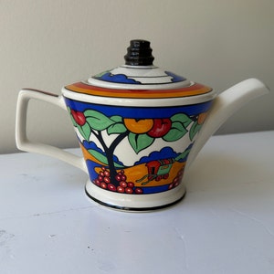 Puede incluir: Una tetera de cer&aacute;mica blanca con asa y tapa negras. La tetera est&aacute; decorada con un dise&ntilde;o colorido con frutas, &aacute;rboles y un carro. La tapa tiene un pomo negro y el pico es blanco.