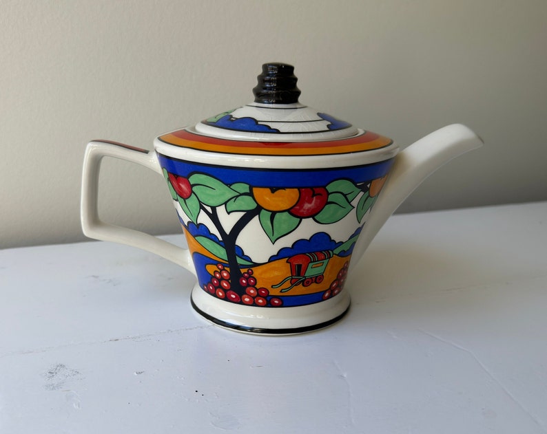 Puede incluir: Una tetera de cer&aacute;mica blanca con asa y tapa negras. La tetera est&aacute; decorada con un dise&ntilde;o colorido con frutas, &aacute;rboles y un carro. La tapa tiene un pomo negro y el pico es blanco.
