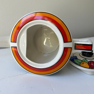 Puede incluir: Tetera de cer&aacute;mica blanca con un dise&ntilde;o circular en naranja, rojo y amarillo. La tapa presenta un patr&oacute;n de frutas y hojas. La tetera tiene un pico y un asa.