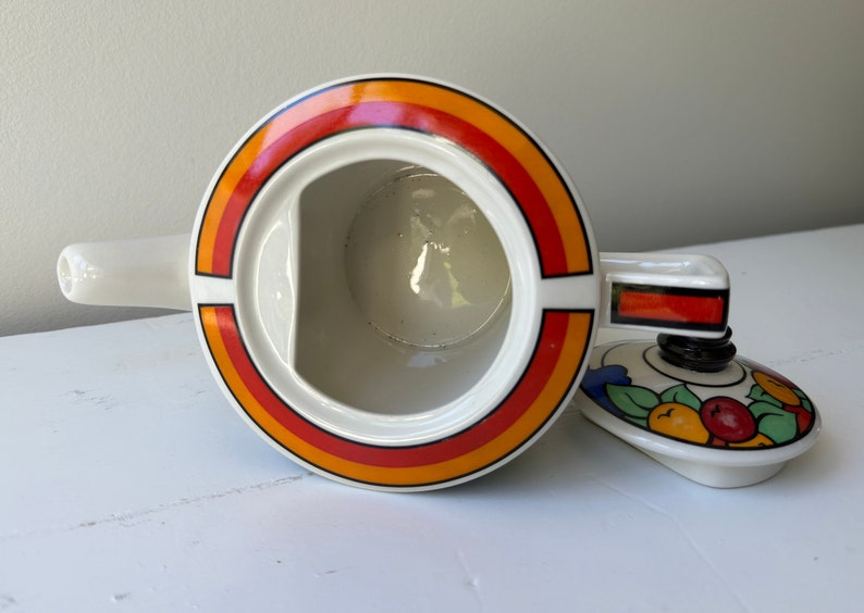 Puede incluir: Tetera de cer&aacute;mica blanca con un dise&ntilde;o circular en naranja, rojo y amarillo. La tapa presenta un patr&oacute;n de frutas y hojas. La tetera tiene un pico y un asa.
