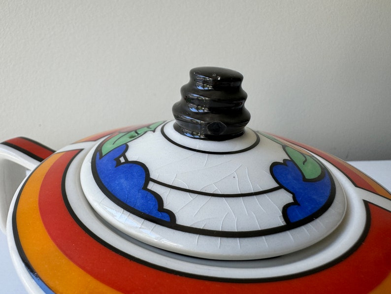 Puede incluir: Primer plano de una tapa de tetera de cer&aacute;mica con un pomo negro. La tapa presenta una superficie blanca con dise&ntilde;os florales azules, verdes y negros. La tetera tiene un patr&oacute;n de arco&iacute;ris de rayas rojas, naranjas y amarillas.