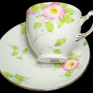 Peut inclure: Une tasse à thé et une soucoupe blanches avec un motif floral. La tasse a une bordure dorée et une anse. La soucoupe a une bordure dorée et une rose rose avec des feuilles vertes. Le fond de la tasse porte le numéro 1935 A.