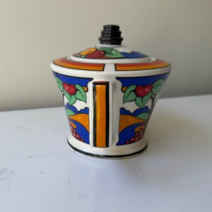 Puede incluir: Un azucarero de cer&aacute;mica blanca con tapa, con un dise&ntilde;o colorido de frutas, hojas y formas geom&eacute;tricas. El azucarero tiene un asa negra y un pomo negro en la tapa. El dise&ntilde;o incluye detalles en naranja, rojo, azul y verde.