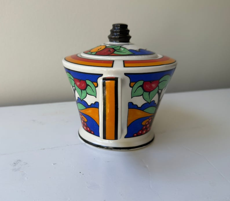 Puede incluir: Un azucarero de cer&aacute;mica blanca con tapa, con un dise&ntilde;o colorido de frutas, hojas y formas geom&eacute;tricas. El azucarero tiene un asa negra y un pomo negro en la tapa. El dise&ntilde;o incluye detalles en naranja, rojo, azul y verde.