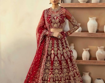 May include: A red and gold embroidered lehenga, a traditional South Asian garment, featuring intricate gold threadwork. The ensemble includes a matching blouse with sheer sleeves and a dupatta. The model is wearing a headpiece and earrings.