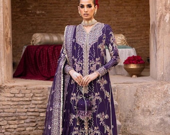 Könnte beinhalten: Ein violettes besticktes Kleid mit goldenen Details und einem passenden Dupatta. Das Kleid hat eine lange, fließende Silhouette und einen hohen Ausschnitt. Der Dupatta ist durchsichtig und hat einen zarten Rand. Das Model trägt goldenen Schmuck und Schuhe.