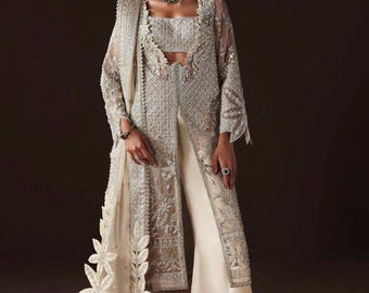 Puede incluir: Conjunto formal marfil adornado. El conjunto incluye una chaqueta larga y transparente con intrincados bordados, un top corto a juego y pantalones de pernera ancha. Una bufanda decorativa con detalles florales y tacones plateados completan el look.