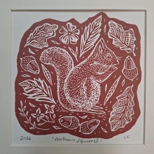 May include: A square linocut print in terracotta red and white featuring a squirrel surrounded by acorns, leaves, and small flowers. The print is titled 'Autumn Squirrel' in handwritten text at the bottom, dated 2026, and signed with the initials cc.