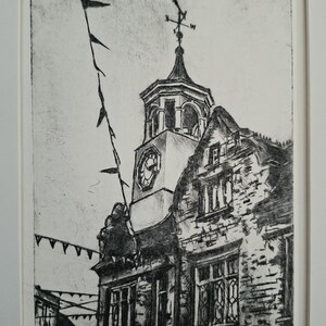 May include: A black and white etching of the Ampthill Clock Tower, featuring a prominent clock face, a weather vane, and decorative bunting hanging from the structure. The artwork is signed and dated 2024, presented in a white mat frame.