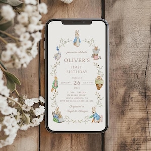 May include: A smartphone displays a first birthday invitation for Oliver. The invitation features illustrations of Peter Rabbit and friends, with text including the date, time, and location of the event. The design is framed by a floral wreath.