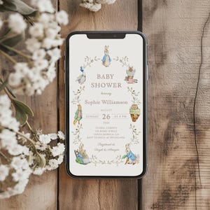 Peut inclure: Un smartphone affiche une invitation à une baby shower. L'invitation présente une couronne florale avec des illustrations de lapins et d'autres animaux. Le texte indique "Baby Shower en l'honneur de Sophie Williamson" et comprend la date, l'heure et le lieu.