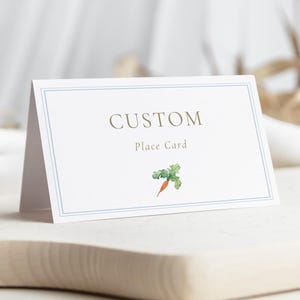 May include: A white place card with a blue border and the word "CUSTOM" in gold lettering. Below the word "CUSTOM" is the text "Place Card" and a watercolor illustration of a carrot. The card is displayed on a wooden surface.