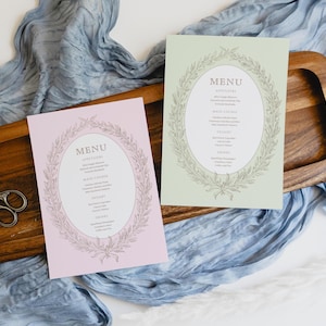 Può includere: Due menu, uno rosa e uno verde, con un design ovale e la parola "MENU" in alto. I menu sono su un vassoio di legno con un paio di forbici e un tessuto blu. I menu hanno un bordo floreale.