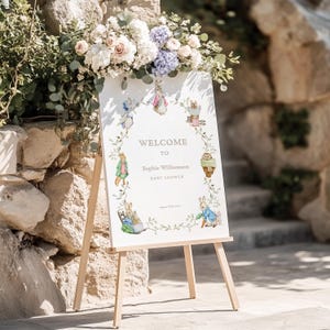 May include: A white welcome sign on a wooden easel, adorned with floral arrangements and illustrations of rabbits. The sign reads "Welcome to Sophie Williamson Baby Shower."