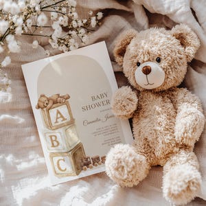 May include: A baby shower invitation with the text "Baby Shower" and the name "Cammelia Jones". The invitation features a watercolor illustration of a teddy bear and alphabet blocks. A plush teddy bear sits next to the invitation.